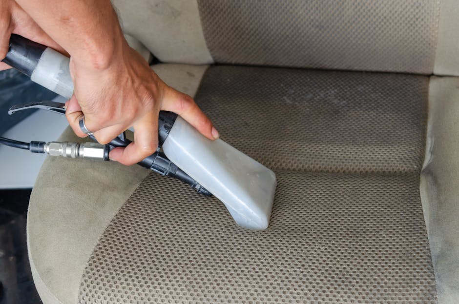 A person using a handheld vacuum cleaner to clean a yellow cushion with a white geometric pattern, placed on a dark fabric sofa in a well-lit room. The vacuum hose is black, and the person's hand is v