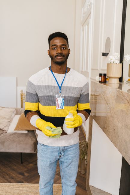 A person wearing white coveralls and yellow gloves is operating a yellow vacuum cleaner with a gray hose attachment, cleaning large gray tiled floors in a modern living room. The room features a gray