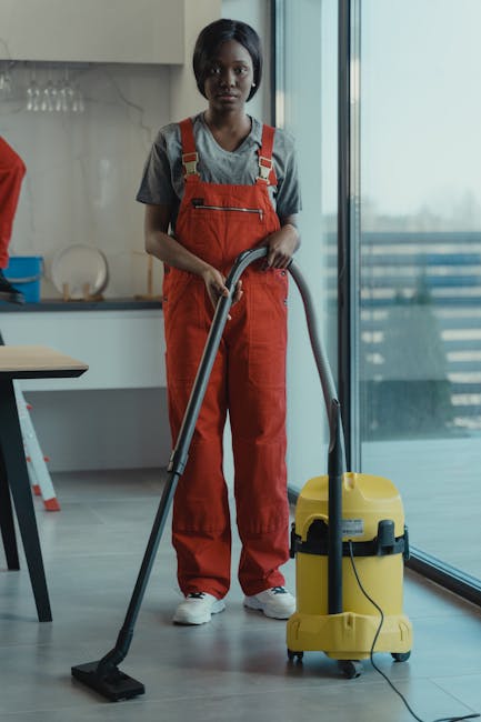 A professional cleaner wearing a yellow uniform and blue gloves stands in a bright, tidy living room, holding a spray bottle of cleaning solution in one hand and a microfiber cloth in the other. The r