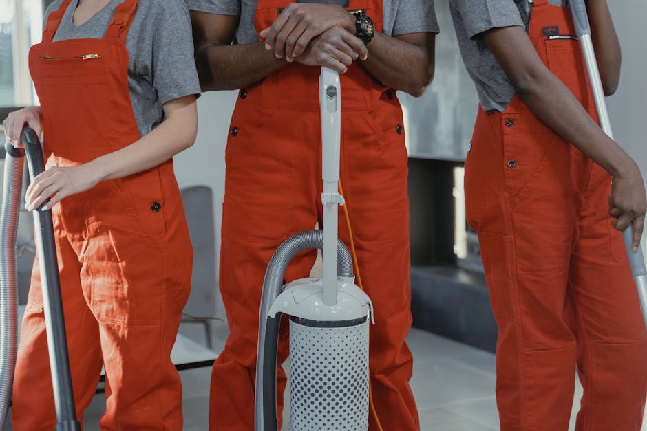 Two professional cleaning technicians dressed in protective work clothing and caps are preparing cleaning equipment outdoors on a paved area. One technician is adjusting or inspecting a portable clean