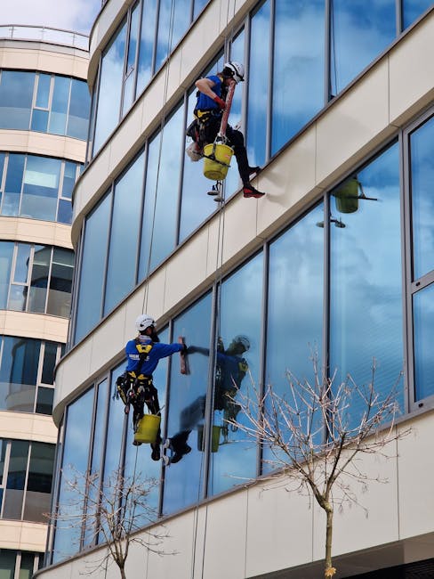 A professional window cleaning operation on a modern high-rise building with a glass facade, featuring a suspended platform with a worker on it, cleaning the large glass panels. The building's exterio